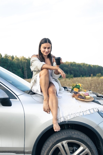 Kostenloses Foto attraktive junge frau sitzt auf einem auto und trinkt kaffee auf einem feld