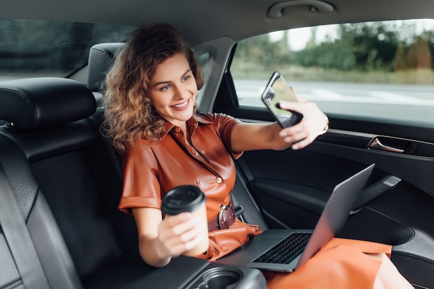 Attraktive junge Geschäftsfrau, die mit einer Tasse Kaffee am Auto arbeitet und das Telefon hält, während sie zur Arbeit geht