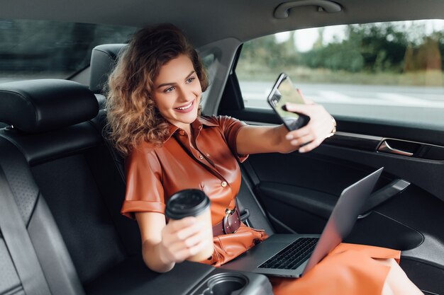 Attraktive junge Geschäftsfrau, die mit einer Tasse Kaffee am Auto arbeitet und das Telefon hält, während sie zur Arbeit geht