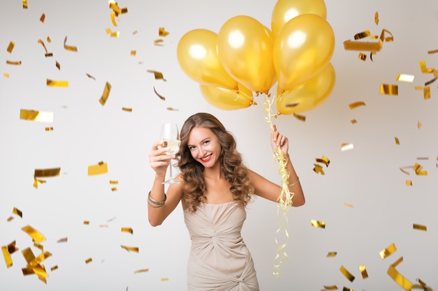 Attraktive junge stilvolle Frau, die neues Jahr feiert, Champagner hält, der Luftballons hält, goldenes Konfetti fliegt, glücklich lächelt, isoliert, Partykleid trägt