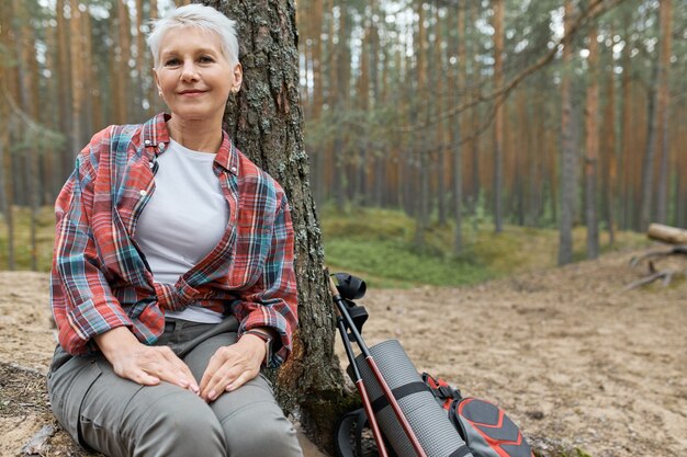 Attraktive kaukasische reife Frau in Aktivkleidung, die unter Baum mit Rucksack neben ihr entspannt, lächelt, beim Wandern im Wald Atem holt, Frieden und Ruhe der wilden Natur genießt