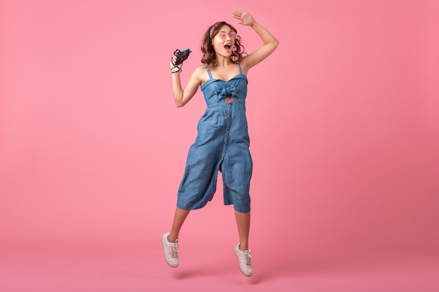 Attraktive lächelnde Frau, die aktiv springt und Foto auf Weinlesekamera trägt, die Denimkleid lokalisiert auf rosa Hintergrund trägt