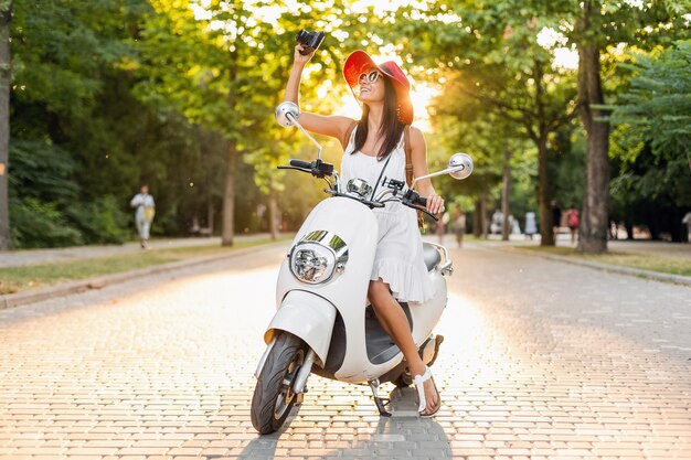 Attraktive lächelnde Frau, die auf Motorrad in der Straße im Sommerart-Outfit reitet, das weißes Kleid und roten Hut trägt, der im Urlaub reist und Fotos auf Vintage-Fotokamera macht
