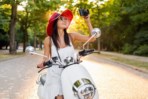 Attraktive lächelnde Frau, die auf Motorrad in der Straße im Sommerart-Outfit reitet, das weißes Kleid und roten Hut trägt, der im Urlaub reist und Fotos auf Vintage-Fotokamera macht