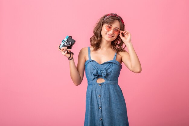 Attraktive lächelnde Frau, die Foto auf Weinlesekamera trägt, die Jeanskleid und Sonnenbrille trägt, lokalisiert auf rosa Hintergrund