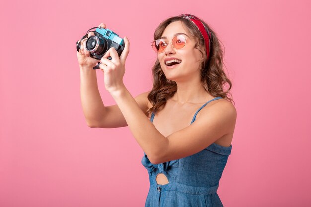 Attraktive lächelnde Frau, die Foto auf Weinlesekamera trägt, die Jeanskleid und Sonnenbrille trägt, lokalisiert auf rosa Hintergrund