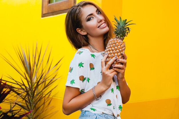 Attraktive lächelnde Frau im Urlaub in der gedruckten T-Shirt-Sommermode, Hände, die Ananas halten