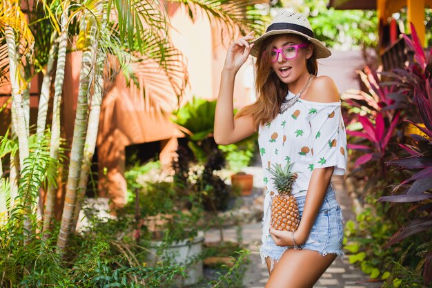 Attraktive lächelnde Frau im Urlaub in der gedruckten T-Shirt-Strohhut-Sommermode, Hände, die Ananas halten