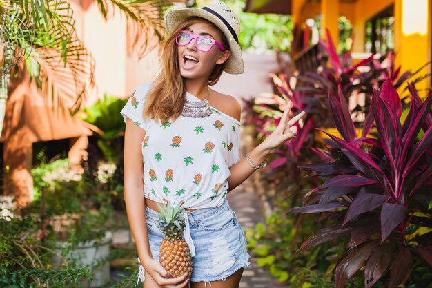 Attraktive lächelnde Frau im Urlaub in der gedruckten T-Shirt-Strohhut-Sommermode, Hände, die Ananas halten