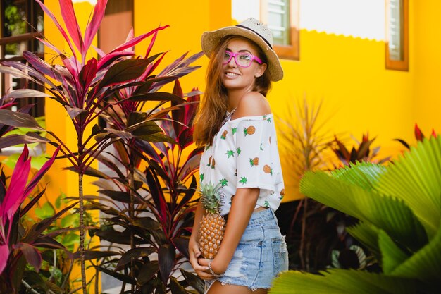 Attraktive lächelnde Frau im Urlaub in der gedruckten T-Shirt-Strohhut-Sommermode, Hände, die Ananas halten