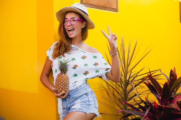 Attraktive lächelnde Frau im Urlaub in der gedruckten T-Shirt-Strohhut-Sommermode, Hände, die Ananas halten