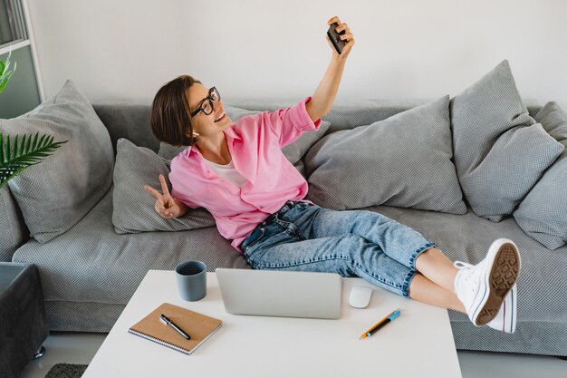 Attraktive lächelnde Frau in rosafarbenem Hemd sitzt entspannt auf dem Sofa zu Hause am Tisch und arbeitet online