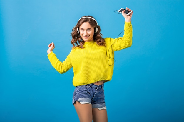Attraktive lächelnde glückliche Frau tanzt, die Musik in den Kopfhörern im stilvollen Hipster-Outfit lokalisiert auf blauem Studiohintergrund hört, der Shorts und gelben Pullover trägt