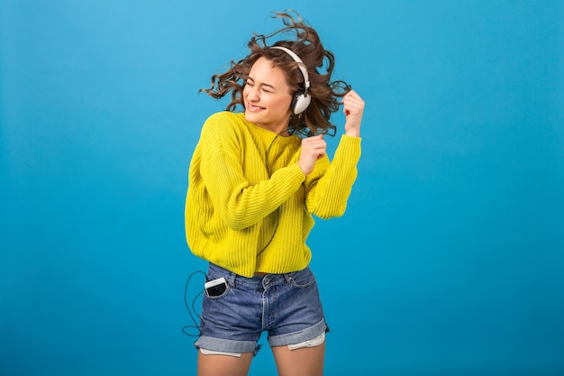 Attraktive lächelnde glückliche Frau tanzt, die Musik in den Kopfhörern im stilvollen Hipster-Outfit lokalisiert auf blauem Studiohintergrund hört, der Shorts und gelben Pullover trägt