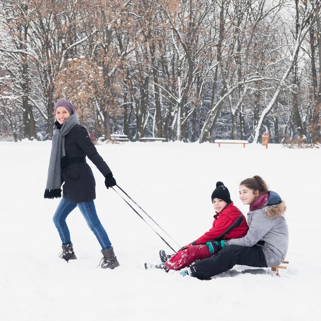 Attraktive lächelnde Mutter, die ihre Kinder auf Schlitten im Winter zieht