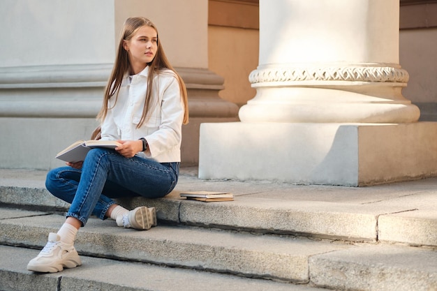 Attraktive lässige Studentin, die nachdenklich mit Buch auf der Universitätstreppe im Freien studiert