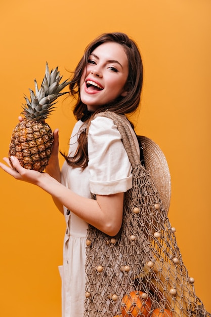 Attraktive langhaarige Frau im weißen Kleid posiert mit gestrickter Einkaufstasche und hält Ananas.