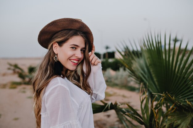 Attraktive langhaarige lachende Frau im trendigen Hut posiert gerne neben grüner Pflanze. Entzückende junge Frau, die weiße Bluse trägt, die in der Wüste geht und lächelt, genießt Urlaub
