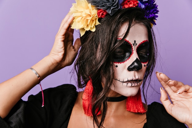 Attraktive lateinamerikanische Dame mit ungewöhnlicher Kunst auf ihrem Gesicht schaut nach unten. Nahaufnahmeporträt der Brünette mit langen roten Ohrringen.