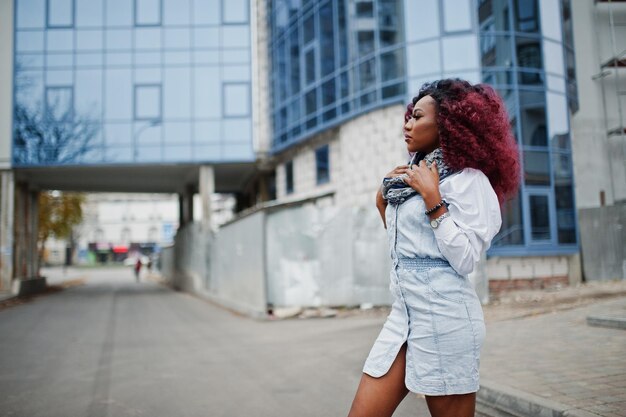 Attraktive lockige Afroamerikanerin im Jeanskleid posierte vor einem modernen mehrstöckigen Gebäude