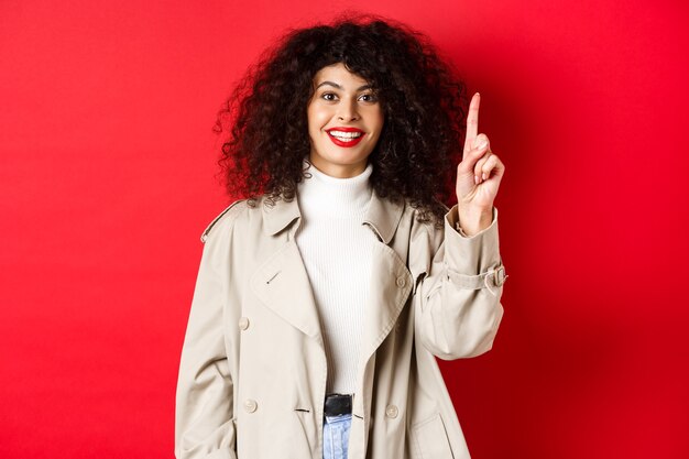 Attraktive moderne Frau mit lockiger Frisur der roten Lippen, die Frühlings-Trenchcoat trägt und mit dem Finger nach oben zeigt ...