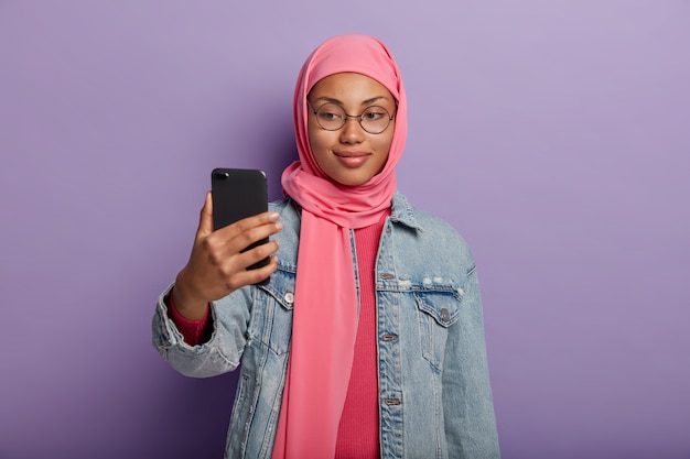 Attraktive muslimische Frau mit kleinem Lächeln, macht Foto von sich selbst über Smartphone, gekleidet in traditioneller Kleidung nach religiösen Überzeugungen.