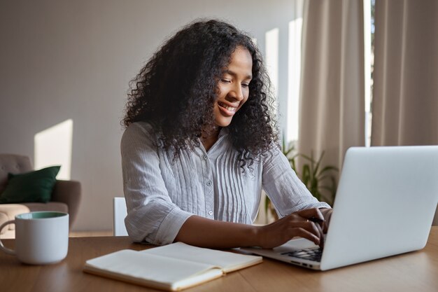 Attraktive positive junge afroamerikanische Freiberuflerin, die aus der Ferne arbeitet, auf einem generischen Laptop tastet, zu Hause mit Heft und Becher auf dem Tisch sitzt, elektronische Nachricht online schreibt und lächelt