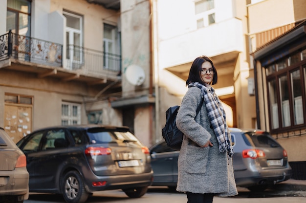 Attraktive positive tragende Gläser des jungen Mädchens in einem Mantel auf dem Hintergrund von Gebäuden auf Autos