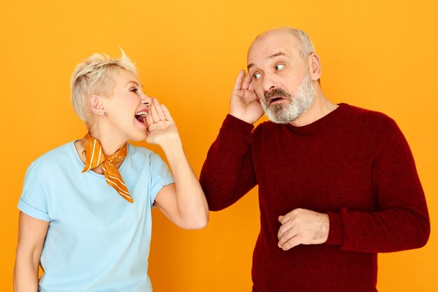 Attraktive reife Frau mit kurzen grauen Haaren, die schreien, während sie sich an ihren Ehemann wendet, der wegen eines Hörproblems die Hand an seinem Ohr hält