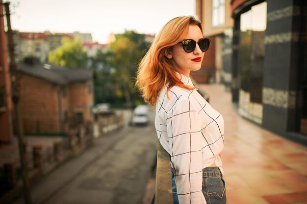 Attraktive rothaarige Frau mit Sonnenbrille trägt eine weiße Bluse und posiert auf der Straße vor einem modernen Gebäude