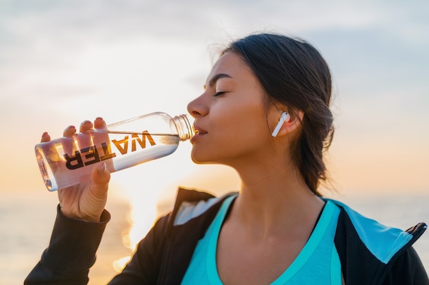 Attraktive schlanke Frau, die Sportübungen am Morgensonnenaufgangstrand in der Sportkleidung, durstiges Trinkwasser in der Flasche, gesunder Lebensstil tut, Musik auf drahtlosen Kopfhörern hörend, glücklich lächelnd macht