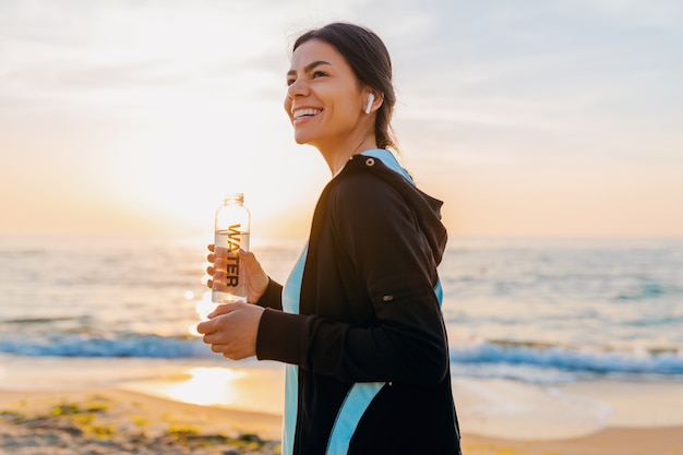 Attraktive schlanke Frau, die Sportübungen am Morgensonnenaufgangstrand in der Sportkleidung, durstiges Trinkwasser in der Flasche, gesunder Lebensstil tut, Musik auf drahtlosen Kopfhörern hörend, glücklich lächelnd macht