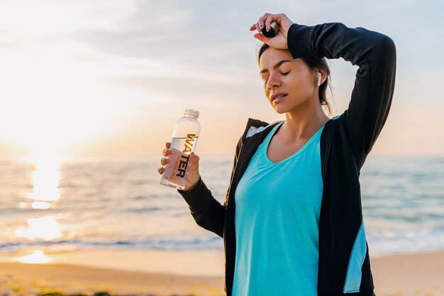 Attraktive schlanke Frau, die Sportübungen am Morgensonnenaufgangstrand in Sportbekleidung, durstiges Trinkwasser in der Flasche, gesunder Lebensstil, Musik hören auf drahtlosen Kopfhörern, heißer Sommertag tut