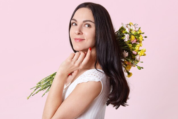 Attraktive schlanke junge Dame hält Blumen auf der Schulter, berührt das Kinn mit den Fingern, steht seitlich und schaut in eine andere Richtung