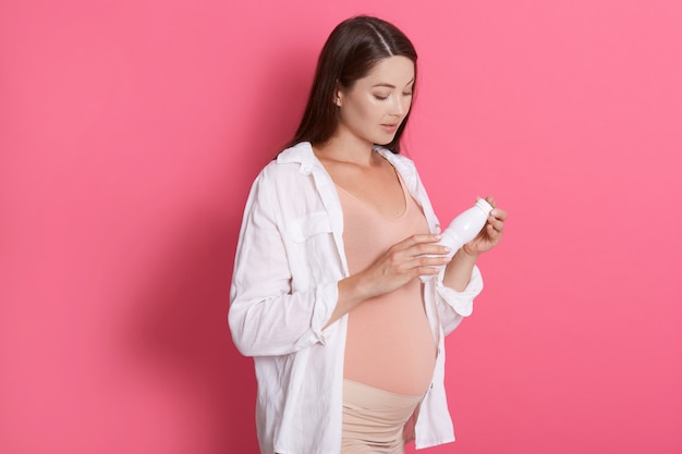 Attraktive schwangere Frau, die Zusammensetzung des Produkts liest, Flasche oder Joghurt oder Milch hält, gegen rosa Wand steht, gesunde Ernährung für zukünftige Mutter und Baby.