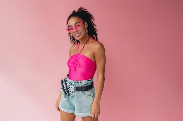 Attraktive schwarze afroamerikanerin im stylischen sommeroutfit, rosafarbenes oberteil und kurze gürteltasche auf rosafarbenem studiohintergrund, sommermodetrend, glücklich lächelndes lockiges haar, sonnenbrillenzubehör