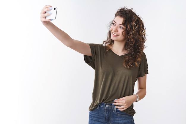 Attraktive, selbstbewusste, lockere Bloggerin, die ein Selfie macht, möchte ein neues Bild posten, einen persönlichen Blog, den Arm ausstrecken, der das Smartphone hält, das in der Nähe des kühlen Hintergrunds posiert, lächelndes Telefondisplay