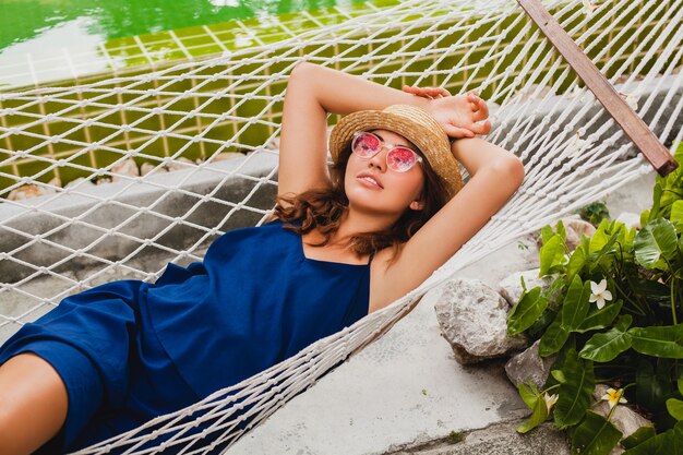 Attraktive sexy junge Frau im blauen Kleid und im Strohhut, die rosa Sonnenbrille tragen, die im Urlaub in der Hängematte liegend entspannen