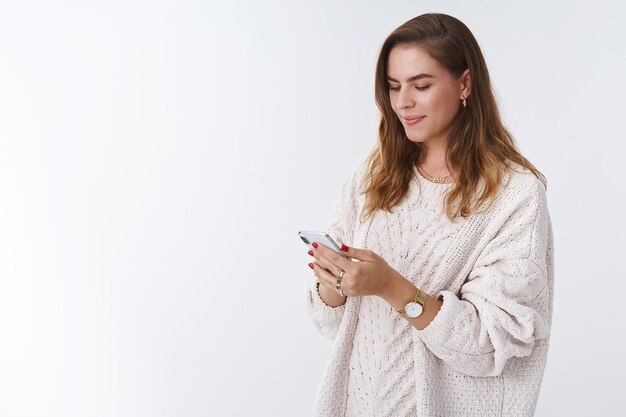Attraktive SMM-Managerin, die eine Webseite verwaltet, die eine Smartphone-Eingabenachricht hält, die lächelnd aussieht, freut sich über einen positiven Handy-Bildschirm, der einen lustigen Videofreund sendet, der auf weißem Hintergrund steht