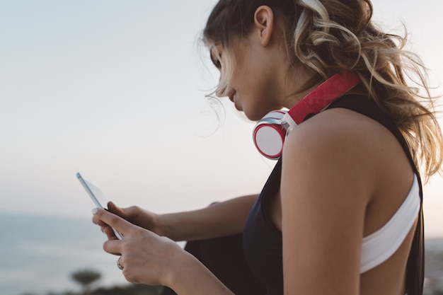 Attraktive Sportlerin des Nahaufnahmeporträts mit Kopfhörern am Hals, langes lockiges Haar, das am Telefon plaudert und Sonnenaufgang am Meer genießt. Fröhliche Stimmung, schöne Sommerzeit.