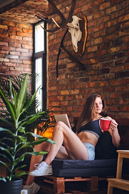 Attraktive sportliche Frau, die Tablet-PCs benutzt und Kaffee im Loft-Innenraum trinkt.