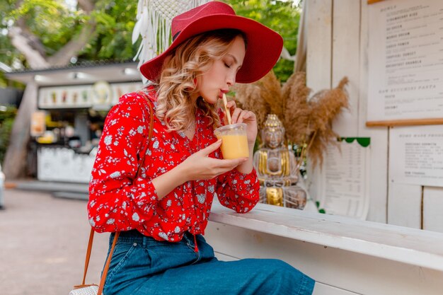 Attraktive stilvolle blonde lächelnde Frau im strohroten Hut und im Sommermode-Outfit der Bluse, die natürlichen Fruchtcocktail-Smoothie trinkt