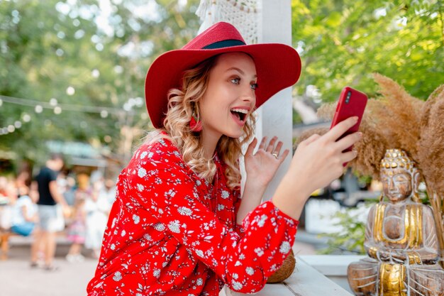 Attraktive stilvolle blonde lächelnde Frau in strohrotem Hut und Bluse Sommermode-Outfit, das unter Verwendung des Smartphone-Cafés hält