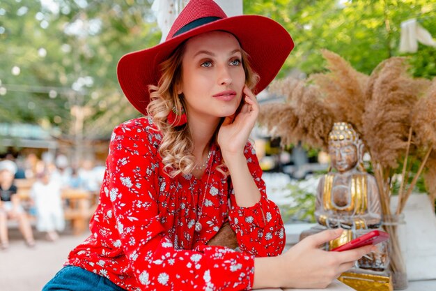 Attraktive stilvolle blonde lächelnde Frau in strohrotem Hut und Bluse Sommermode-Outfit, das unter Verwendung des Smartphone-Cafés hält