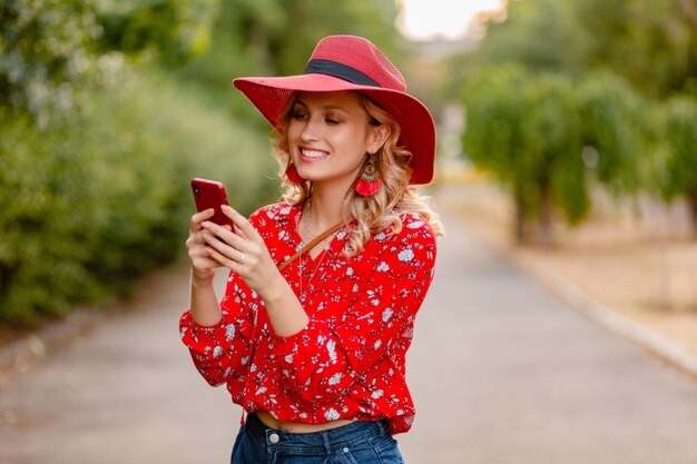 Attraktive stilvolle blonde lächelnde Frau in strohrotem Hut und Bluse Sommermode-Outfit unter Verwendung des Telefons