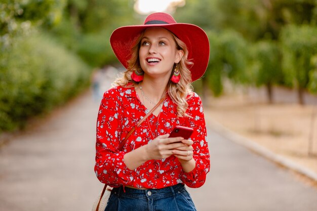 Attraktive stilvolle blonde lächelnde Frau in strohrotem Hut und Bluse Sommermode-Outfit unter Verwendung des Telefons