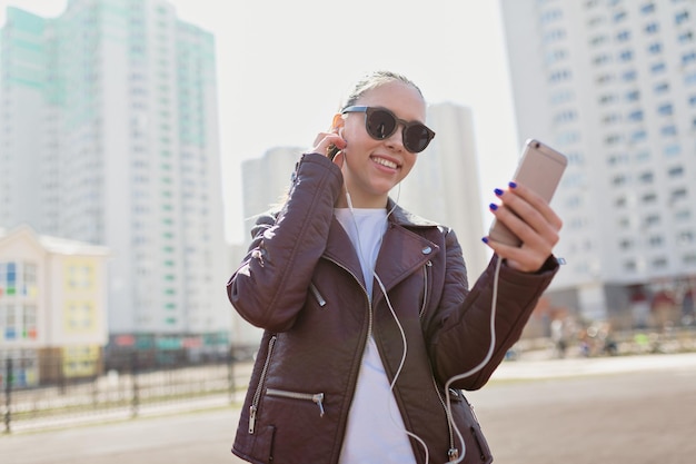 Attraktive stilvolle europäische frau mit dunklem haar, sonnenbrille und jacke trägt smartphone und hört musik un kopfhörer auf stadthintergrund