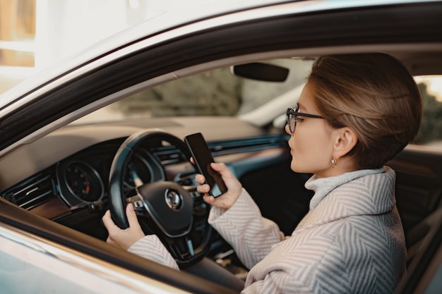 Attraktive stilvolle Frau, die im Auto sitzt, gekleidet in Mantelwinterstil und Brille mit Smartphone