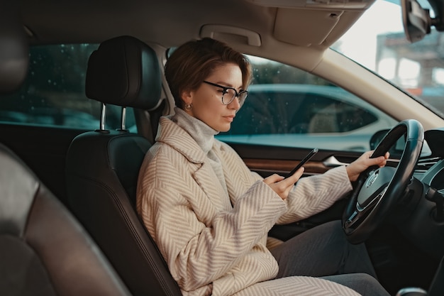 Attraktive stilvolle Frau, die im Auto sitzt, gekleidet in Mantelwinterstil und Brille mit Smartphone