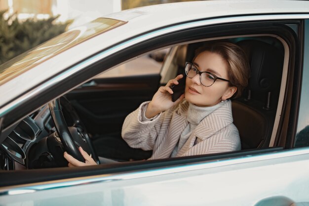 Attraktive stilvolle Frau, die im Auto sitzt, gekleidet in Mantelwinterstil und Brille mit Smartphone